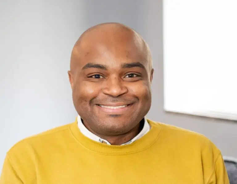 Daniel Plunkett, a Black male therapist, sits in a cozy therapy office wearing a mustard yellow sweater, exuding warmth and confidence.