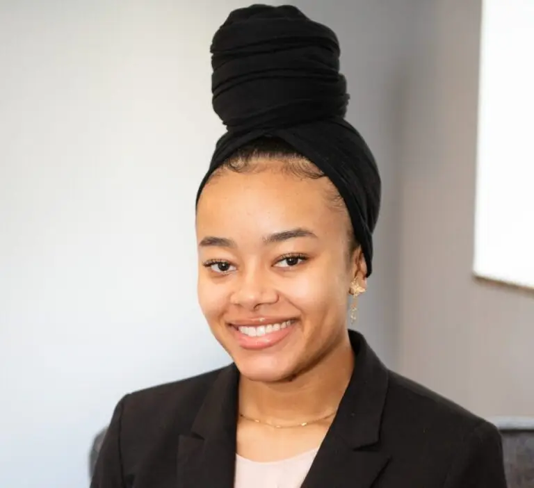 Kalila F., an intern therapist, sits on a gray couch in an office, wearing a black blazer over a soft pink blouse. Her smile is inviting and professional.