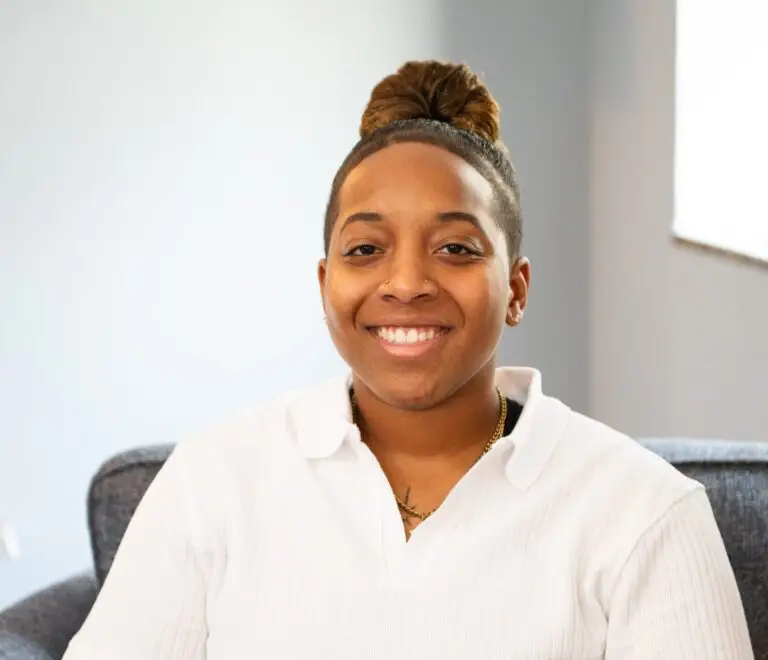 Matiah, an intern therapist, sits comfortably on a gray couch in a well-lit office, wearing a white polo and brown pants. She smiles warmly with a relaxed posture.