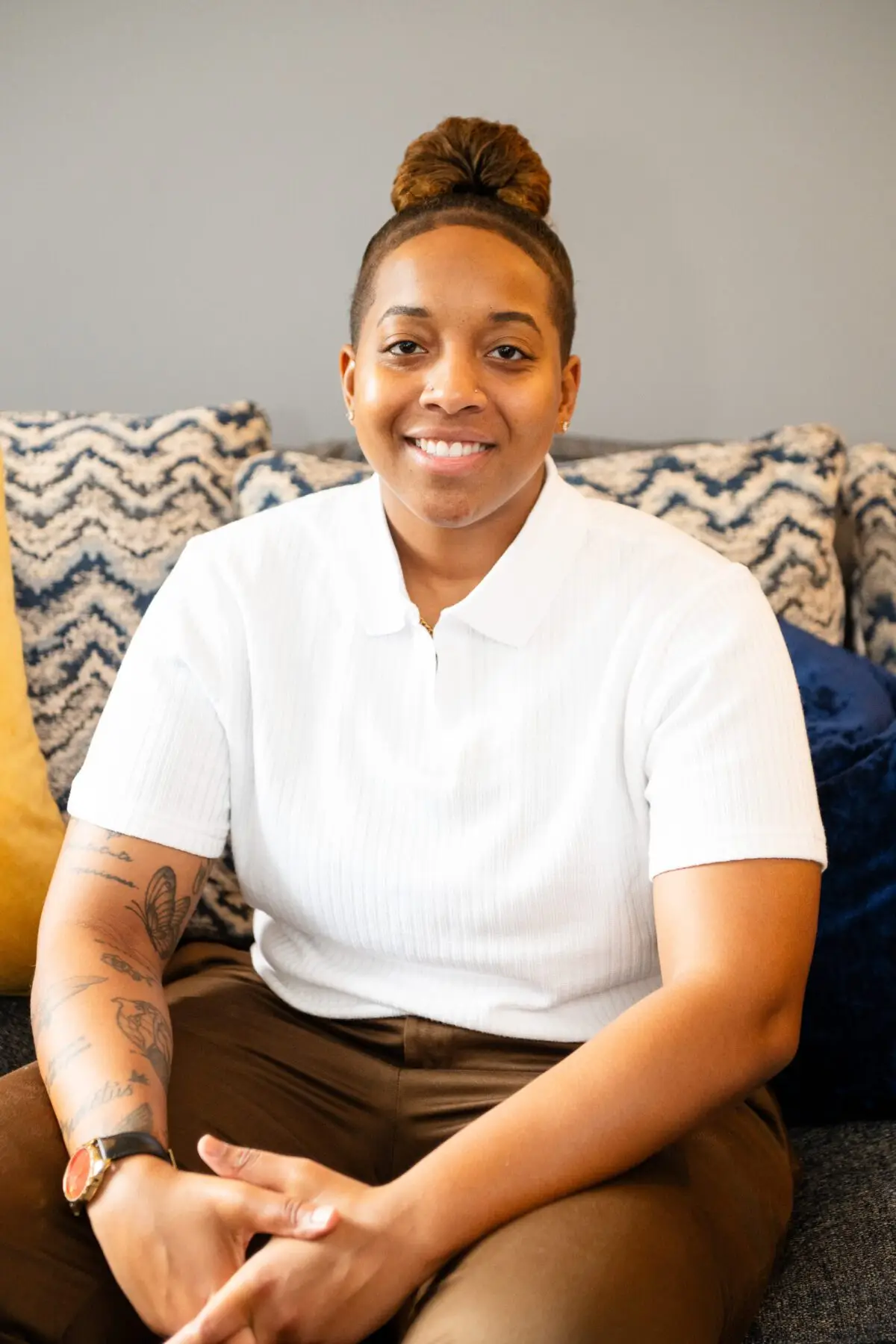 Matiah, an intern therapist, sits on a stylish couch with patterned pillows, wearing a white polo shirt and brown pants. Her smile conveys warmth and professionalism.