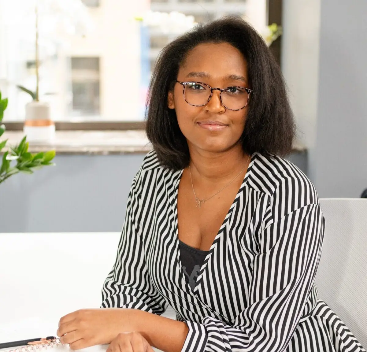 Nyia Hawkins, an intern therapist, sits at a white desk with an open notebook, pen in hand. She wears a striped blouse and blue jeans, looking engaged and ready to support clients.