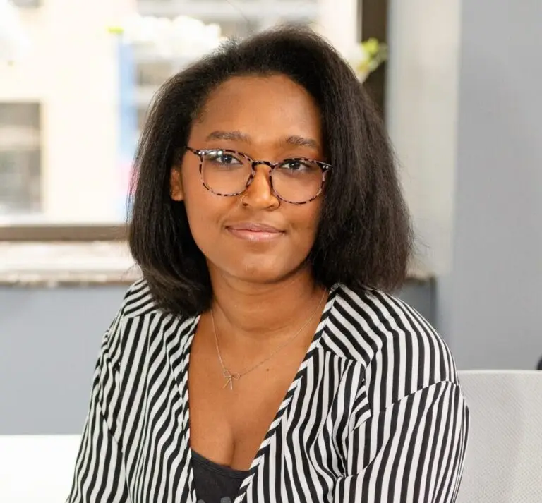 Nyia Hawkins, an intern therapist, sits at a white desk with an open notebook, pen in hand. She wears a striped blouse and blue jeans, looking engaged and ready to support clients.