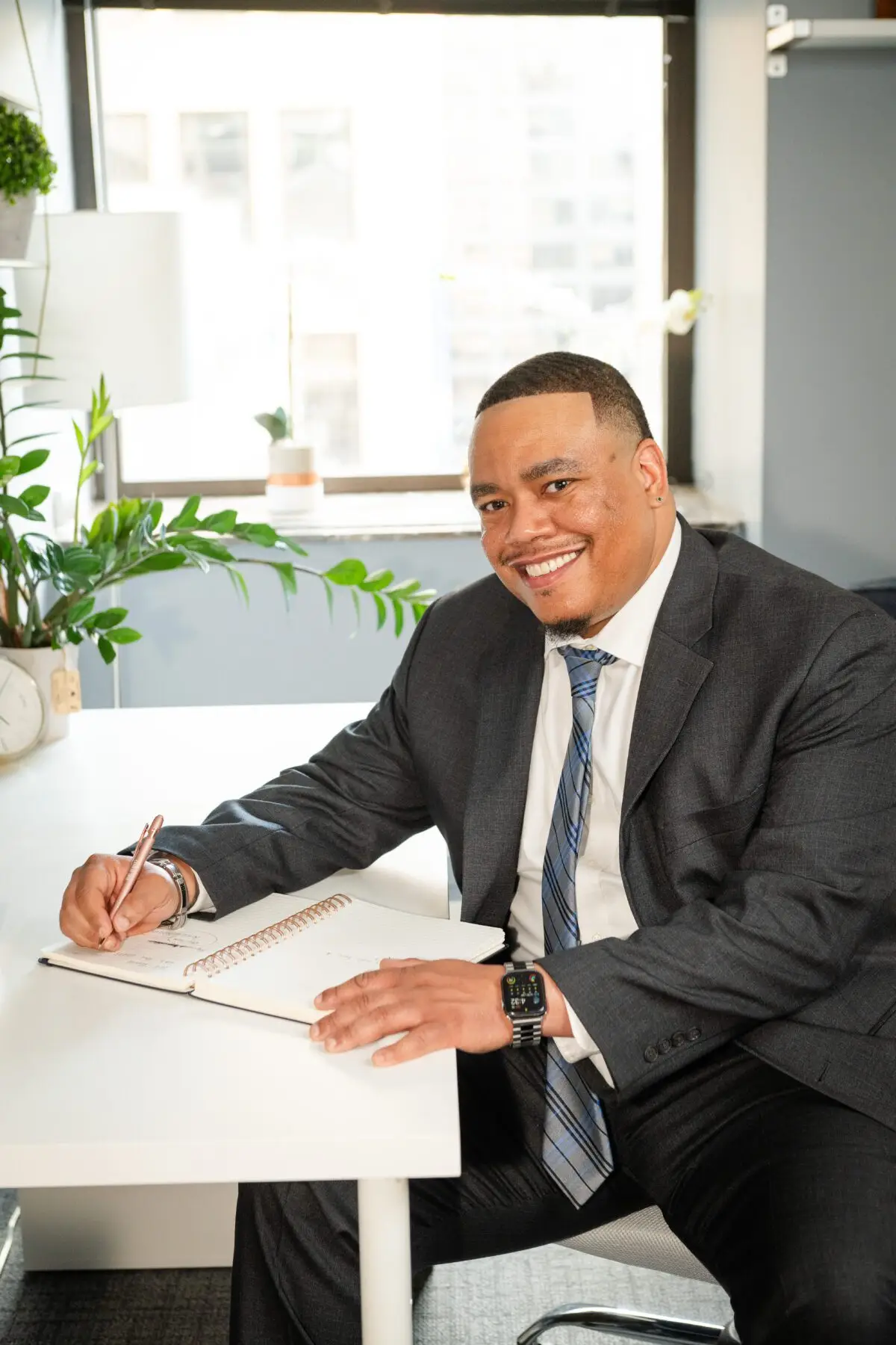 Dr. Perrin Greene, a Black male therapist, sits at his desk in a professional suit, smiling warmly as he writes in a notebook.