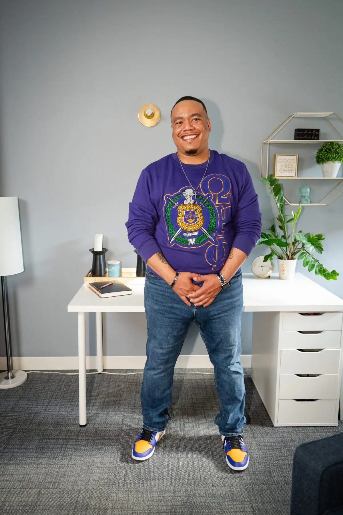 Dr. Perrin Greene, a Black male therapist, stands confidently in his office, wearing a purple Omega Psi Phi sweater and jeans.