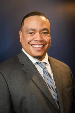 Dr. Perrin Greene, a Black male therapist, dressed in a professional suit, smiling confidently against a deep blue background.