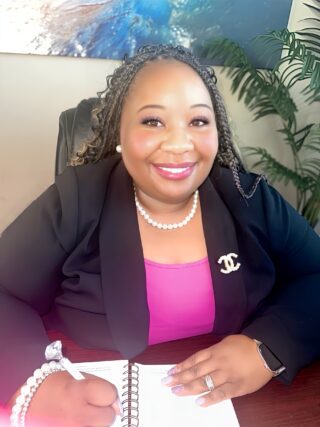 Erica Butler sitting at a desk, smiling confidently in a professional outfit with a stylish brooch and pearls, exuding warmth and professionalism.