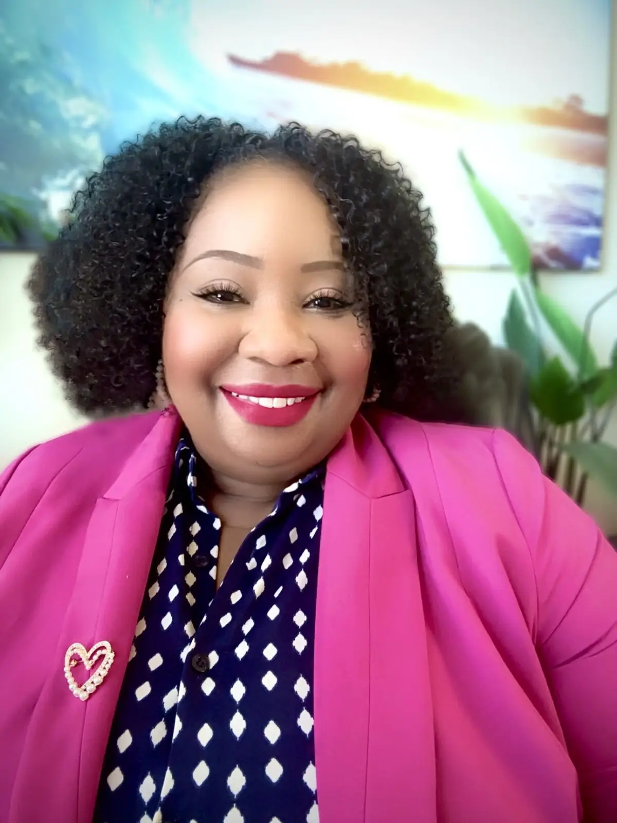 Erica Butler smiling in a vibrant pink blazer and patterned shirt, seated in a cozy office with a backdrop of plants and serene artwork.